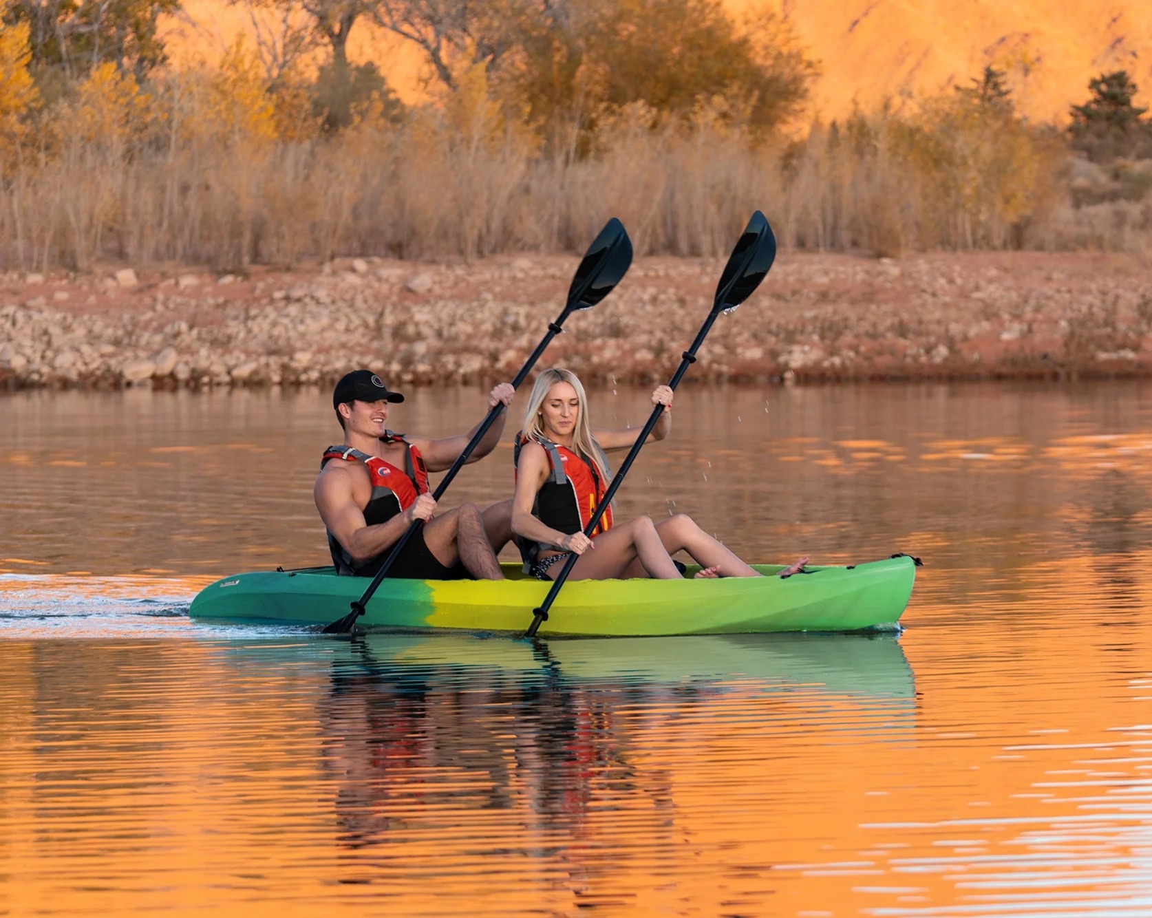 A man and woman paddle a green kayak together on a calm lake at sunset, with autumn trees in the background.