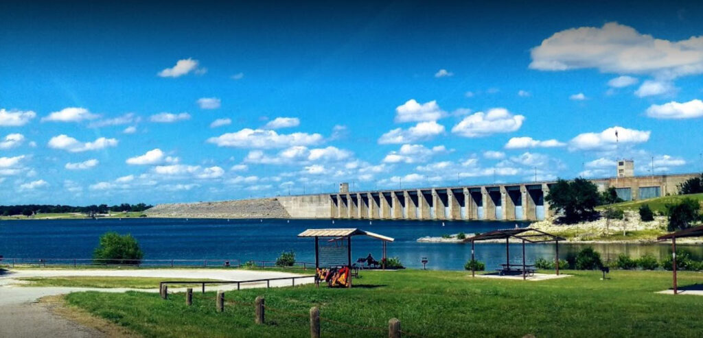 Whitneytxdam A large dam stretches across a wide blue reservoir under a bright sky dotted with fluffy clouds, with grassy picnic shelters and a walking path in the foreground.