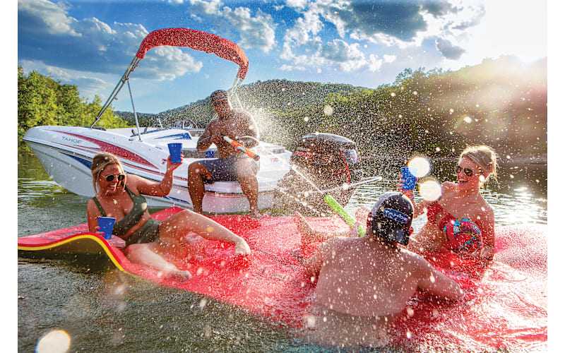 Four people relax on a floating mat in the water, splashing and holding blue cups, while another person sits on a nearby boat under a sunny sky.