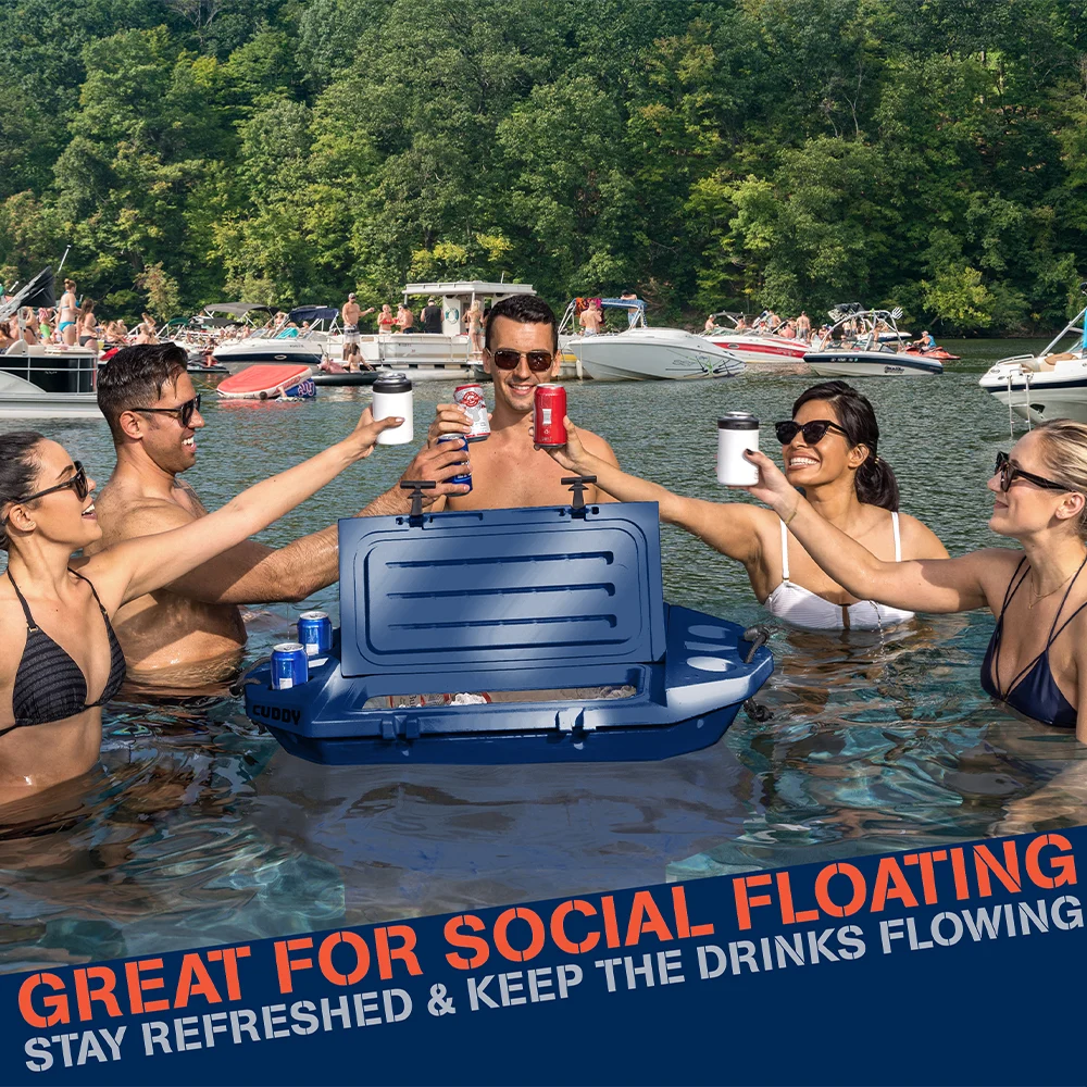 Five people in swimsuits cheer with drinks around a Go Sports Cuddy floating cooler in a lake, surrounded by boats and lush trees under a sunny sky.